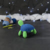 Amigurumi représentant une tortue verte à carapace bleue, vue de dos orientée vers la gauche, posée sur un fond noir avec des pelotes bleues, un crochet et une guirlande lumineuse en arrière-plan.