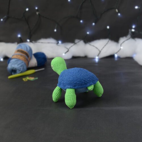 Amigurumi représentant une tortue verte à carapace bleue, vue de dos orientée vers la gauche, posée sur un fond noir avec des pelotes bleues, un crochet et une guirlande lumineuse en arrière-plan.