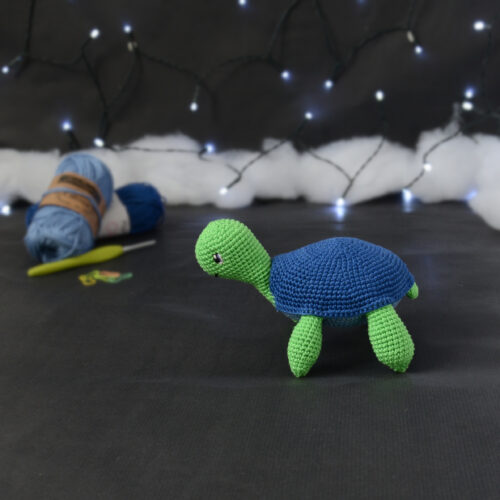 Amigurumi représentant une tortue verte à carapace bleue, de profil orientée vers la gauche, posée sur un fond noir avec des pelotes bleues, un crochet et une guirlande lumineuse en arrière-plan.