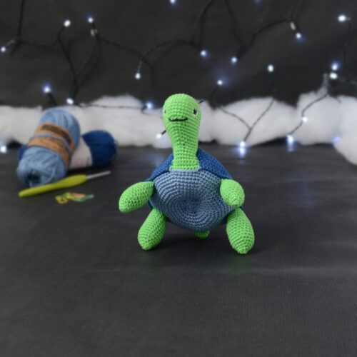 Amigurumi représentant une tortue verte à carapace bleue, vue de face, dressée sur ses pattes arrières, posée sur un fond noir avec des pelotes bleues, un crochet et une guirlande lumineuse en arrière-plan.