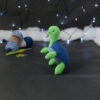 Amigurumi représentant une tortue verte à carapace bleue, dressée sur ses pattes arrières, vue de côté orientée vers la gauche, posée sur un fond noir avec des pelotes bleues, un crochet et une guirlande lumineuse en arrière-plan.