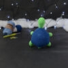 Amigurumi représentant une tortue verte à carapace bleue, dressée sur ses pattes arrières, vue de dos, posée sur un fond noir avec des pelotes bleues, un crochet et une guirlande lumineuse en arrière-plan.