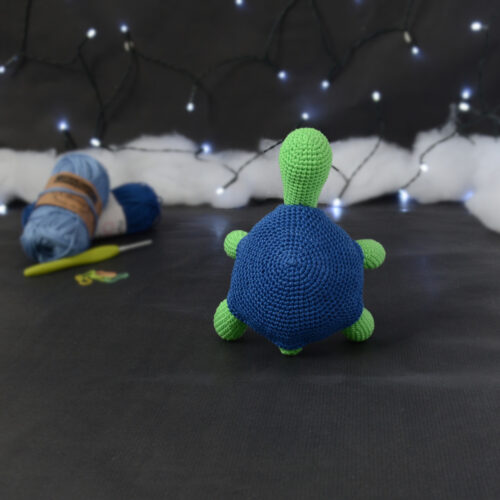 Amigurumi représentant une tortue verte à carapace bleue, dressée sur ses pattes arrières, vue de dos, posée sur un fond noir avec des pelotes bleues, un crochet et une guirlande lumineuse en arrière-plan.