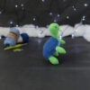 Amigurumi représentant une tortue verte à carapace bleue, dressée sur ses pattes arrières, vue de côté orientée vers la droite, posée sur un fond noir avec des pelotes bleues, un crochet et une guirlande lumineuse en arrière-plan.