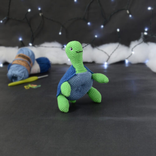 Amigurumi représentant une tortue verte à carapace bleue, dressée sur ses pattes arrières, vue de côté orientée vers la droite, posée sur un fond noir avec des pelotes bleues, un crochet et une guirlande lumineuse en arrière-plan.
