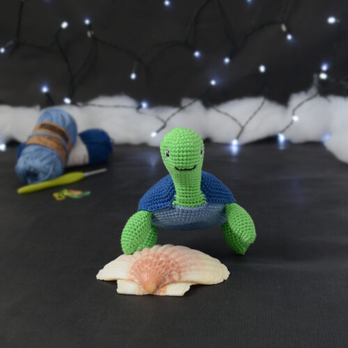 Amigurumi représentant une tortue verte à carapace bleue, vue de face, posée sur un coquillage, sur un fond noir avec des pelotes bleues, un crochet et une guirlande lumineuse en arrière-plan.