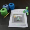Sur un fond foncé est posé une page du patron de Géraldine la tortue. Sur le côté droit de cette page se trouve la tortue crochetée en vert et bleu, dressée sur ses pattes arrières. Dans le coin supérieur gauche de la page il y a trois pelotes de coton vert, bleu et bleu clair avec un crochet jaune.