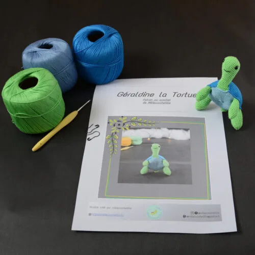 Sur un fond foncé est posé une page du patron de Géraldine la tortue. Sur le côté droit de cette page se trouve la tortue crochetée en vert et bleu, dressée sur ses pattes arrières. Dans le coin supérieur gauche de la page il y a trois pelotes de coton vert, bleu et bleu clair avec un crochet jaune.