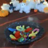 Un lot de bonbons au crochet dans une panière grise foncé au crochet dans une ambiance d'Halloween. Le tout est posé sur un tissu orange, avec en arrière-plan des décorations d'Halloween crochetées : citrouilles, fantômes et chauve-souris sur un fond de nuages blancs.