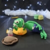 Grenouille amigurumi verte allongée sur une bouée canard jaune, portant un short violet, avec un chapeau et une boisson crochetés posés à côté, sur fond sombre décoré de guirlande lumineuse.