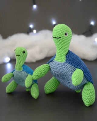 Deux tortues crochetées, l'une grande et l'autre petite, de couleur vert et bleu sont posées côte à côté sur un fond gris foncé. Il y a un nuage de ouate en fond ainsi qu'une guirlande lumineuse.