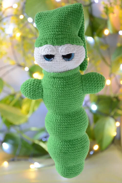 Luciole au crochet, de couleur verte avec un visage clair et de grands yeux. Elle porte un grand bonnet retombant vers l'arrière. Elle est photographiée devant un fond flou végétalisé et avec des guirlandes lumineuses.