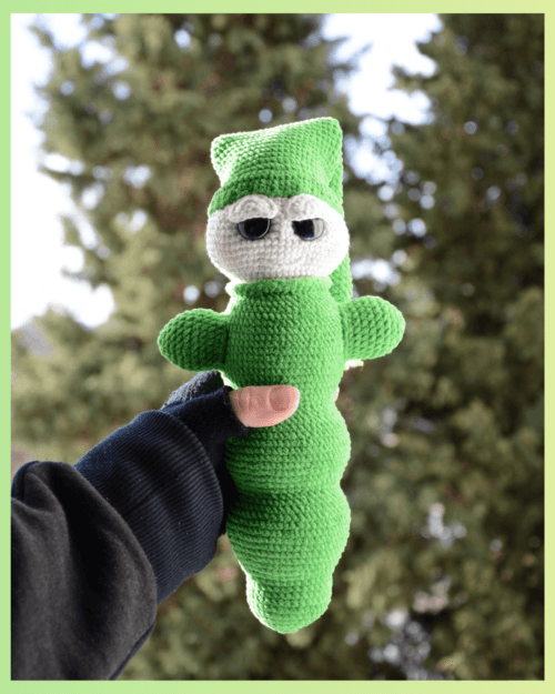 Luciole au crochet, de couleur verte avec un visage clair et de grands yeux. Elle porte un bonnet et est tenue dans une main, photographiée en extérieur devant un arrière-plan naturel flou.