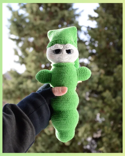 Luciole au crochet, de couleur verte avec un visage clair et de grands yeux. Elle porte un bonnet et est tenue dans une main, photographiée en extérieur devant un arrière-plan naturel flou.