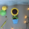 Amigurumi représentant un tournesol dans un pot bleu avec une tige verte et trois feuilles. La fleur a de grands pétales jaunes et un cœur marron foncé avec un visage souriant brodé avec des yeux rieurs fermés. La photo est prise de dessus, il est posé sur une surface grise avec une guirlande lumineuse sur le côté droit et à l'avant-plan. Sur le côté gauche, il y a des pelotes de coton verte et jaune un crochet ainsi que des anneaux marqueurs. À l'arrière-plan, on distingue un voilage bleuté.
