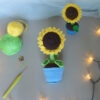 Amigurumi représentant un tournesol dans un pot bleu avec une tige verte et trois feuilles. La fleur a de grands pétales jaunes et un cœur marron foncé avec un visage souriant brodé avec des yeux rieurs fermés. Il y a un autre tournesol plus petit, posé sur le côté gauche. La photo est prise de dessus, il est posé sur une surface grise avec une guirlande lumineuse sur le côté droit et à l'avant-plan. Sur le côté gauche, il y a des pelotes de coton verte et jaune un crochet ainsi que des anneaux marqueurs. À l'arrière-plan, on distingue un voilage bleuté.