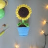 Amigurumi représentant un tournesol dans un pot bleu avec une tige verte et trois feuilles. La fleur a de grands pétales jaunes et un cœur marron foncé avec un visage souriant brodé avec des yeux rieurs fermés. La photo est prise de dessus, il est posé sur une surface grise avec une guirlande lumineuse sur le côté droit et à l'avant-plan. Sur le côté gauche, on distingue des pelotes de coton verte et jaune un crochet ainsi que des anneaux marqueurs.