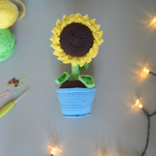 Amigurumi représentant un tournesol dans un pot bleu avec une tige verte et trois feuilles. La fleur a de grands pétales jaunes et un cœur marron foncé avec un visage souriant brodé avec des yeux rieurs fermés. La photo est prise de dessus, il est posé sur une surface grise avec une guirlande lumineuse sur le côté droit et à l'avant-plan. Sur le côté gauche, on distingue des pelotes de coton verte et jaune un crochet ainsi que des anneaux marqueurs.