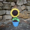 Un tournesol crocheté placé sur un rocher. Il est dans un pot bleu, avec une tige verte et un cœur brun entouré de pétales jaunes. Il a un visage souriant brodé avec des yeux rieurs fermés. À l'arrière-plan, un mur de pierres.