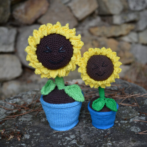 Deux tournesols crochetés de tailles différentes, placés côte à côte sur un rocher. Chaque fleur est dans un pot bleu, avec une tige verte et un cœur brun entouré de pétales jaunes. Les deux tournesols affichent un visage souriant brodé avec des yeux rieurs fermés. À l'arrière-plan, un mur de pierres.