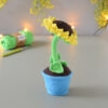 Amigurumi représentant un tournesol dans un pot bleu avec une tige verte et trois feuilles. Il est orienté la fleur levé vers le haut. La fleur a de grands pétales jaunes et un cœur marron foncé. Il est posé sur une surface grise avec en fond une guirlande lumineuse et des fleurs jaunes. En arrière-plan flouté, on distingue une pelote de coton verte ainsi qu'un crochet.