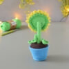 Amigurumi représentant un tournesol dans un pot bleu avec une tige verte et trois feuilles. Il est présenté de dos. La fleur a de grands pétales jaunes. Il est posé sur une surface grise avec en fond une guirlande lumineuse et des fleurs jaunes. En arrière-plan flouté, on distingue une pelote de coton verte ainsi qu'un crochet.