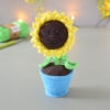 Amigurumi représentant un tournesol dans un pot bleu avec une tige verte et trois feuilles. La fleur a de grands pétales jaunes et un cœur marron foncé avec un visage souriant brodé avec des yeux rieurs fermés. Il est posé sur une surface grise avec en fond une guirlande lumineuse et des fleurs jaunes. En arrière-plan flouté, on distingue une pelote de coton verte ainsi qu'un crochet.