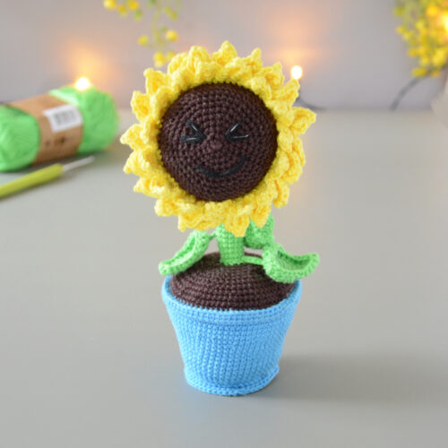 Amigurumi représentant un tournesol dans un pot bleu avec une tige verte et trois feuilles. La fleur a de grands pétales jaunes et un cœur marron foncé avec un visage souriant brodé avec des yeux rieurs fermés. Il est posé sur une surface grise avec en fond une guirlande lumineuse et des fleurs jaunes. En arrière-plan flouté, on distingue une pelote de coton verte ainsi qu'un crochet.