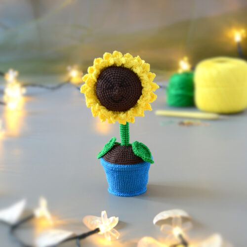 Amigurumi représentant un tournesol dans un pot bleu avec une tige verte et deux feuilles. La fleur a de grands pétales jaunes et un cœur marron foncé. Il est posé sur une surface grise, entouré de guirlandes lumineuses en forme de fleurs. En arrière-plan flouté, on distingue des pelotes de coton jaune et vert ainsi qu'un crochet et des anneaux marqueurs.