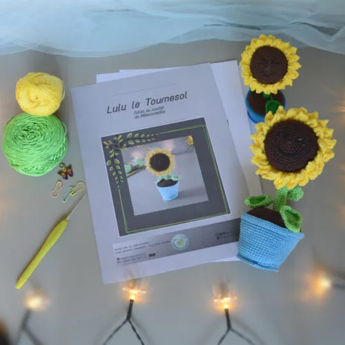 Des pages du patron de lulu le tournesol sont posés sur un fond gris. Sur le côté droit de ces pages se trouve 2 versions crochetées de Lulu le tournesol, une plus grande couchée et l'autre plus petite. L'un des tournesol est posé couché dans son pot, l'autre est debout dans son pot. Les 2 ont le visage orientés vers le haut, semblant nous sourire. À l'avant-plan se trouve une guirlande lumineuse.