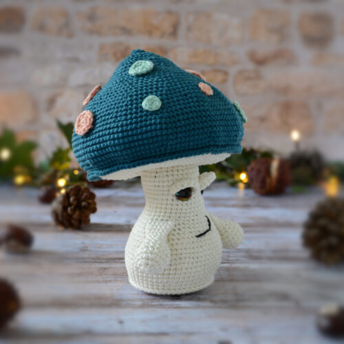 Champignon au crochet, avec un chapeau bleu pétrole décoré de spots rose et vert d'eau. Il est photographié tourné vers le côté droit.
