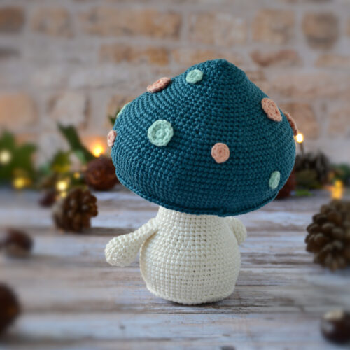 Champignon au crochet, avec un chapeau bleu pétrole décoré de spots rose et vert d'eau. Il est photographié tourné de dos.