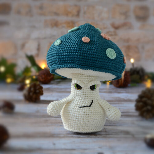 Champignon au crochet, avec un chapeau bleu pétrole décoré de spots rose et vert d'eau. Il est photographié de face, et ses paupières lui donne un air un peu mesquin.