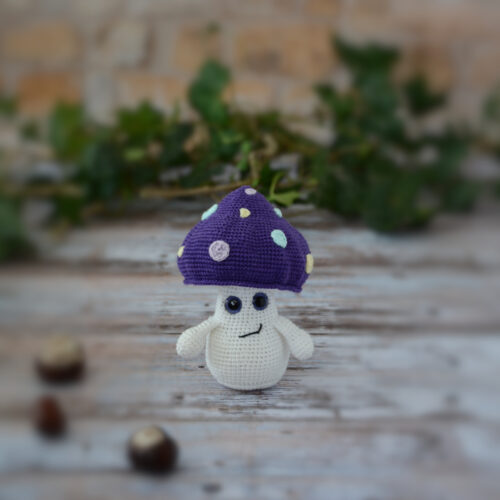 Petit champignon au crochet, avec un chapeau violet décoré de spots colorés. Il est photographié de face, sans paupières.