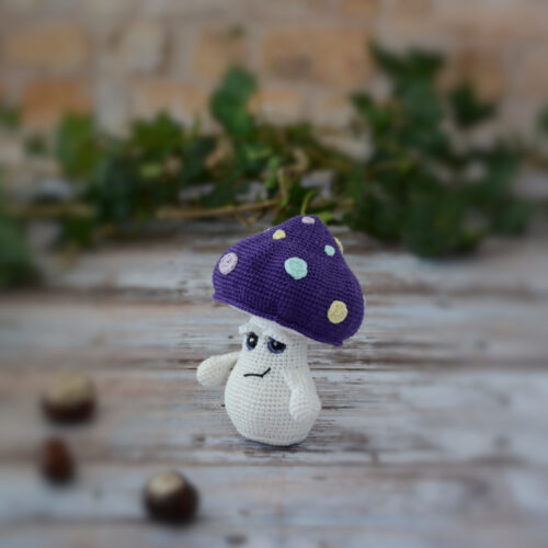 Petit Ottie champignon au crochet, avec un chapeau violet décoré de spots colorés. Il est tourné vers le côté gauche, révélant mieux ainsi ses paupières