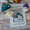 Des pages du patron d'Ottie le champignon sont posées sur une table en bois. Ces pages sont entourées d'un côté et de l'autre de deux versions du champignon, une grande sur la gauche et une plus petite sur la droite. Le grand champignon a un chapeau bleu pétrole et des pois spots rose et vert d'eau. Le petit lui a un chapeau violet décoré de spots coloré. En fond il y a quelques feuilles automnales ainsi qu'un marron. Une guirlande lumineuse entoure le tout.