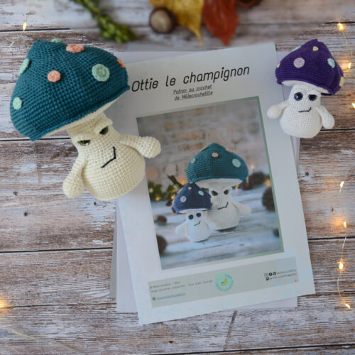 Des pages du patron d'Ottie le champignon sont posées sur une table en bois. Ces pages sont entourées d'un côté et de l'autre de deux versions du champignon, une grande sur la gauche et une plus petite sur la droite. Le grand champignon a un chapeau bleu pétrole et des pois spots rose et vert d'eau. Le petit lui a un chapeau violet décoré de spots coloré. En fond il y a quelques feuilles automnales ainsi qu'un marron. Une guirlande lumineuse entoure le tout.