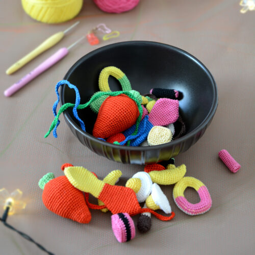 Un bol noir rempli de bonbons crochetés colorés avec notamment deux gros en forme de fraises rouges, un œuf au plat, des réglisses, des bananes et autres. Certains de ces bonbons sont disposés devant le bol. En arrière-plan, on aperçoit des pelotes de coton rose et jaune, des crochets ainsi qu'une guirlande lumineuse.