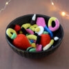 Un bol noir rempli de bonbons crochetés colorés avec notamment deux gros en forme de fraises rouges, un œuf au plat, des réglisses, des bananes et autres. En arrière-plan, on aperçoit une guirlande lumineuse.
