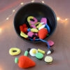Un bol noir rempli de bonbons crochetés colorés avec notamment deux gros en forme de fraises rouges, des œufs au plat, des réglisses, des bananes et autres. Plusieurs de ces bonbons sont devant le bol comme tombés de celui-ci. En arrière-plan, on aperçoit une guirlande lumineuse.