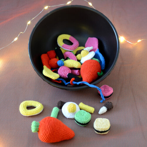 Un bol noir rempli de bonbons crochetés colorés avec notamment deux gros en forme de fraises rouges, des œufs au plat, des réglisses, des bananes et autres. Plusieurs de ces bonbons sont devant le bol comme tombés de celui-ci. En arrière-plan, on aperçoit une guirlande lumineuse.