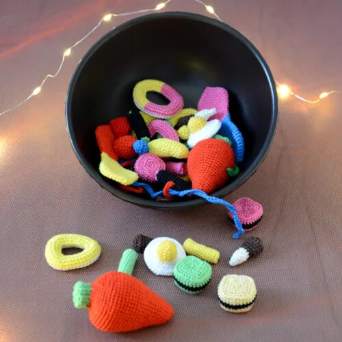 Un bol noir rempli de bonbons crochetés colorés avec notamment deux gros en forme de fraises rouges, des œufs au plat, des réglisses, des bananes et autres. Plusieurs de ces bonbons sont devant le bol comme tombés de celui-ci. En arrière-plan, on aperçoit une guirlande lumineuse.