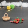 sur un fond gris foncé, sur la gauche il y a un panier crocheté en topaze, et sur sa droite se trouvent des fruits rouges miniatures : fraises, cerises et baies variées (framboises, mûres,...). À l'arrière plan, sur la gauche il y a 2 pelotes jaune et verte et un crochet. En fond est disposé un nuage de ouate et une guirlande lumineuse.