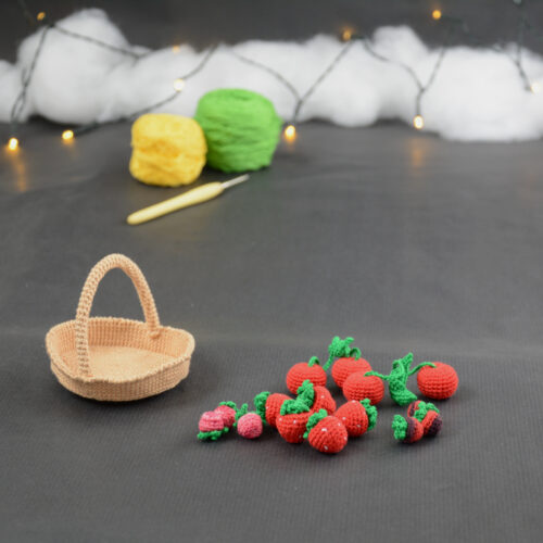 sur un fond gris foncé, sur la gauche il y a un panier crocheté en topaze, et sur sa droite se trouvent des fruits rouges miniatures : fraises, cerises et baies variées (framboises, mûres,...). À l'arrière plan, sur la gauche il y a 2 pelotes jaune et verte et un crochet. En fond est disposé un nuage de ouate et une guirlande lumineuse.