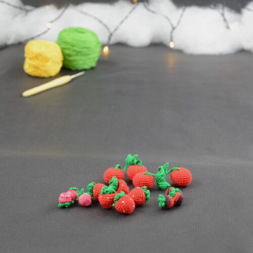 sur un fond gris foncé sont disposés des fruits rouges miniatures crochetés : fraises, cerises et baies variées (framboises, mûres,...). À l'arrière plan, sur la gauche il y a 2 pelotes jaune et verte et un crochet. En fond est disposé un nuage de ouate et une guirlande lumineuse.