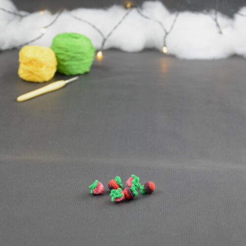 sur un fond gris foncé sont disposés des baies miniatures crochetés : framboises, mûres,... À l'arrière plan, sur la gauche il y a 2 pelotes jaune et verte et un crochet. En fond est disposé un nuage de ouate et une guirlande lumineuse.