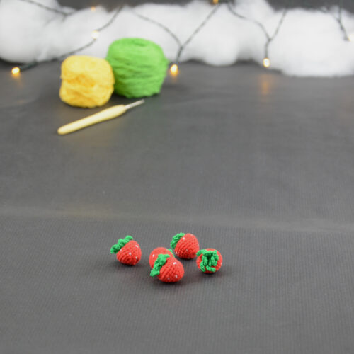 sur un fond gris foncé sont disposés des fraises miniatures crochetés. À l'arrière plan, sur la gauche il y a 2 pelotes jaune et verte et un crochet. En fond est disposé un nuage de ouate et une guirlande lumineuse.