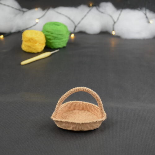 sur un fond gris foncé est disposé un panier miniature crocheté en topaze. À l'arrière plan, sur la gauche il y a 2 pelotes jaune et verte et un crochet. En fond est disposé un nuage de ouate et une guirlande lumineuse.