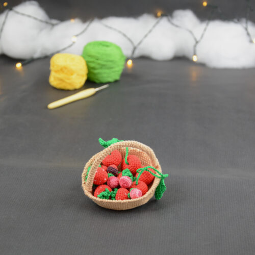 sur un fond gris foncé, il y a un panier crocheté en topaze garni de fruits rouges miniatures : fraises, cerises et baies variées (framboises, mûres,...). À l'arrière plan, sur la gauche il y a 2 pelotes jaune et verte et un crochet. En fond est disposé un nuage de ouate et une guirlande lumineuse.