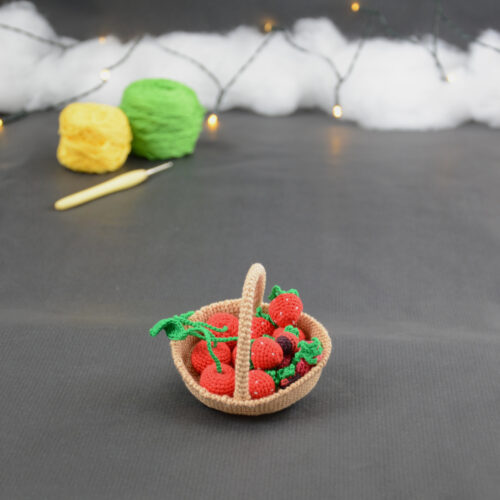 sur un fond gris foncé, il y a un panier crocheté en topaze garni de fruits rouges miniatures : fraises, cerises et baies variées (framboises, mûres,...). À l'arrière plan, sur la gauche il y a 2 pelotes jaune et verte et un crochet. En fond est disposé un nuage de ouate et une guirlande lumineuse.